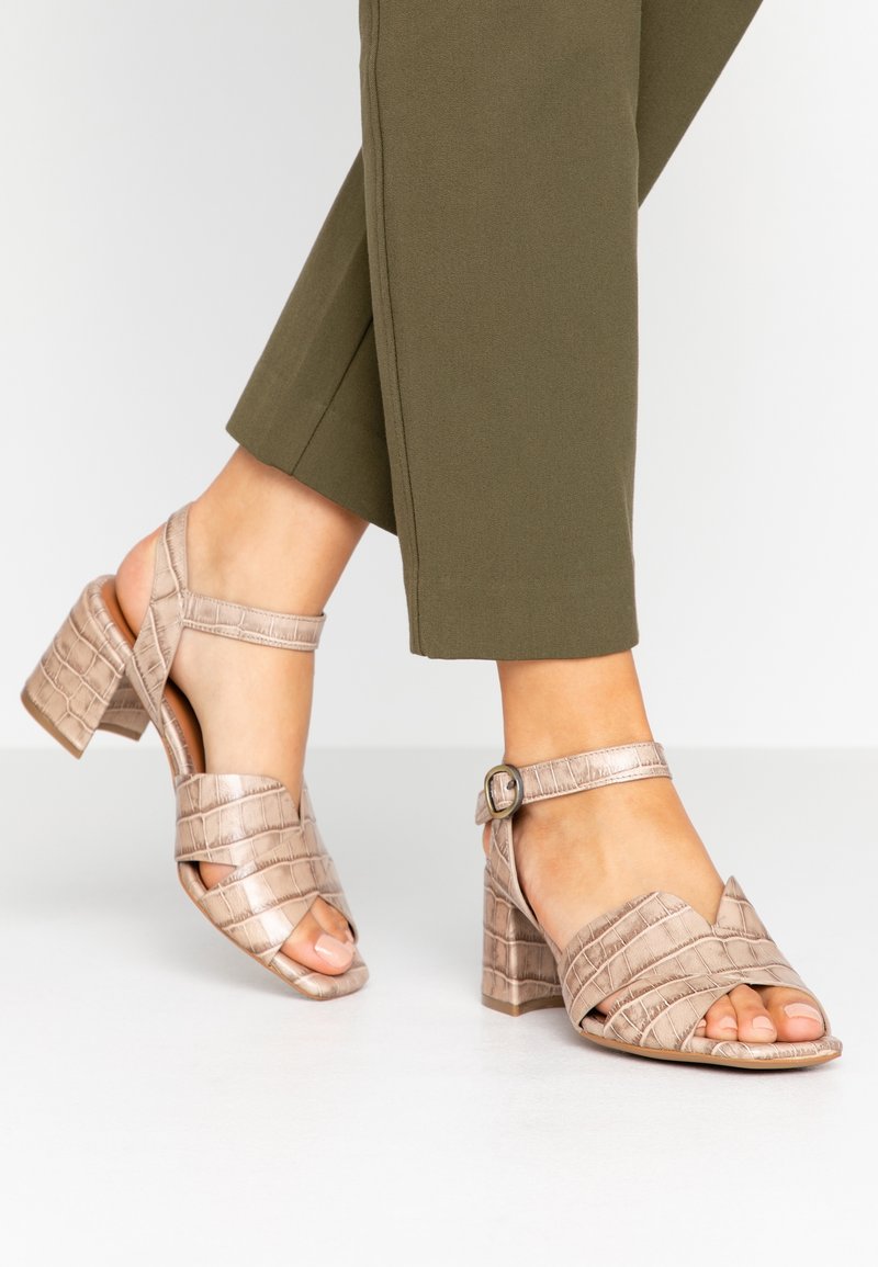 Pieds portant des sandales beiges à talons blocs texturés croco avec brides à la cheville et pantalon court vert olive sur un sol blanc.