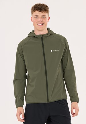 Ung mand smilende iført en grøn lynlåshættejakke med "Virtus" logo og sorte bukser mod en ensartet lys baggrund.
