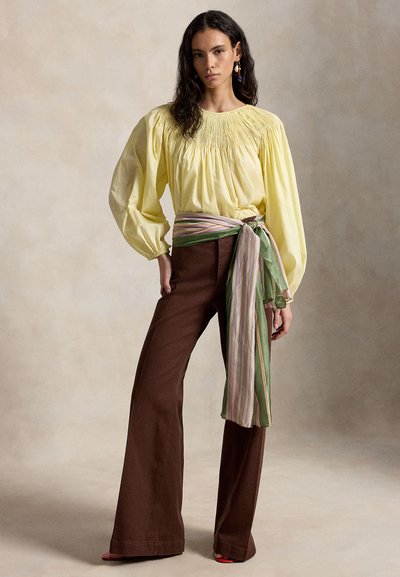 Mujer vistiendo una blusa de manga larga de color amarillo pálido con fruncidos, pantalones marrones de pierna ancha y una bufanda a rayas verde y rosa atada a la cintura.
