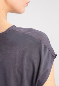 Šedá blůza s hladkým povrchem a krátkými rukávy, se průhledným překrytím na ramenech. Jednoduchý design s volným střihem.