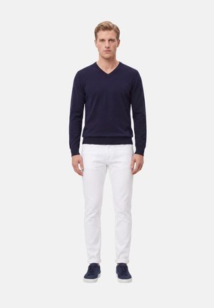 Uomo biondo in piedi di fronte, indossa un maglione blu navy a scollo a V, pantaloni bianchi e sneaker blu navy su uno sfondo bianco uniforme.