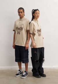Beige polo skjorter med brune detaljer, med grafiske trykk og avslappet passform. Modellen har på seg mørke denimshorts og svarte ripped jeans.