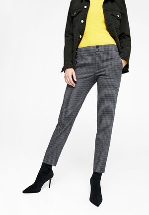 Woman wearing black jacket, yellow sweater, gray checkered pants, and black high-heeled ankle boots standing with one hand in pocket.