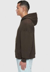 Sweat à capuche marron avec une coupe décontractée, de grandes poches et des poignets côtelés. Le tissu semble doux avec une texture lisse.