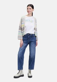 Strickjacke mit mehrfarbigen Streifen, weißes T-Shirt mit dem Text "OCEAN", blaue Jeans und weiße klobige Schnürstiefel.