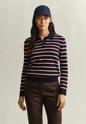 Femme portant une casquette bleu marine et un polo rayé bleu marine à manches longues avec un pantalon marron foncé, debout devant un fond uni.