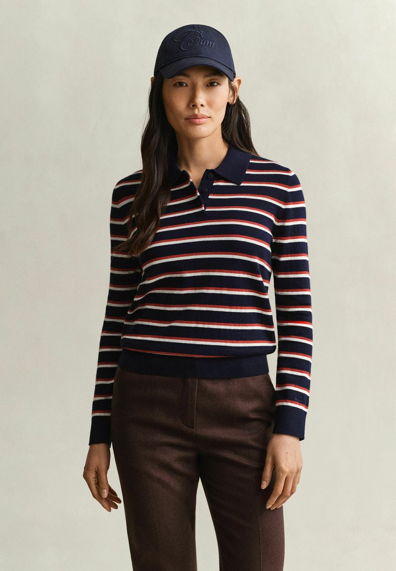 Femme portant une casquette bleu marine et un polo rayé bleu marine à manches longues avec un pantalon marron foncé, debout devant un fond uni.