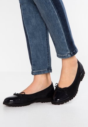 Woman wearing dark blue jeans and black ballet flats with shiny toe caps and small bows, standing on a light floor.