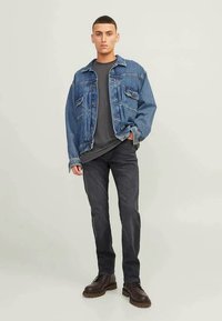 Jaqueta de ganga, azul clara com detalhes nos bolsos, usada sobre uma t-shirt cinza escuro. Jeans pretos e botas castanhas com atacadores completam o visual.