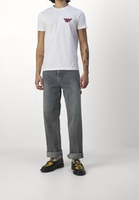 White short-sleeve t-shirt featuring a red and black graphic on the chest, paired with light gray loose-fit jeans and patterned black shoes.