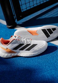 Λευκά αθλητικά παπούτσια με μαύρες ρίγες, πορτοκαλί λεπτομέρειες, υφασμάτινη επένδυση με υφή, καουτσούκ σόλα με σχέδιο ψαροκόκαλου, σχεδιασμένα για απόδοση.