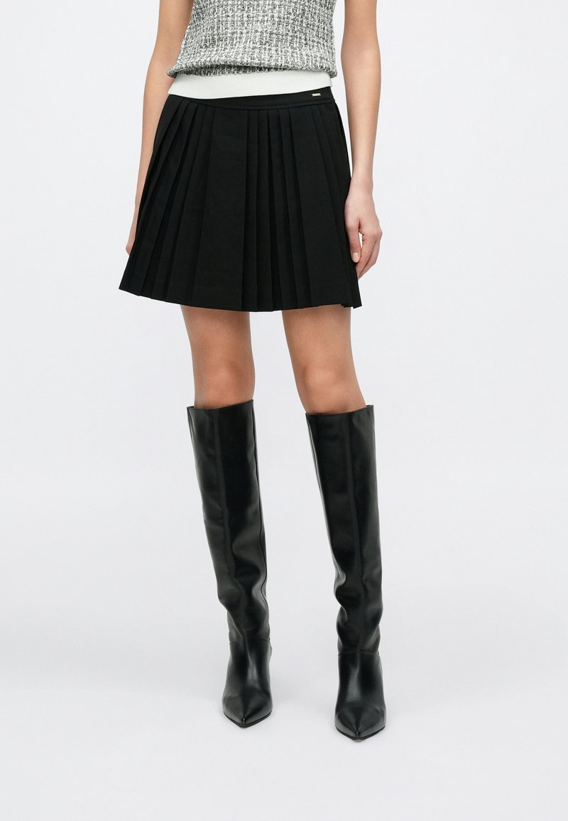 Femme portant une mini-jupe plissée noire, un top gris sans manches et des bottes en cuir noires montant jusqu'aux genoux, debout sur un fond blanc.