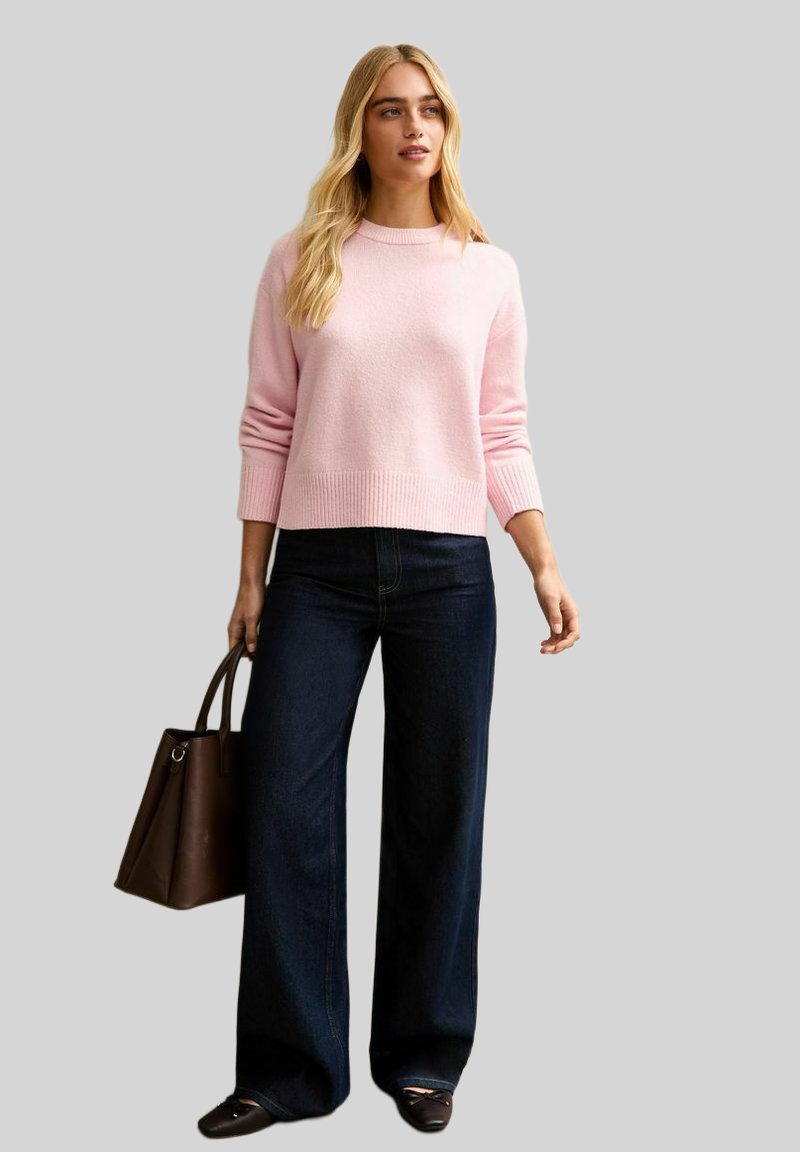 Femme aux cheveux blonds longs portant un pull rose clair, un jean large foncé, des ballerines noires, et tenant un sac à main marron.