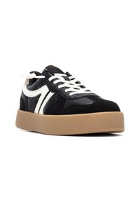 Zapatilla negra hecha de tejido texturizado y ante, con acentos blancos, cordones planos y una suela gruesa beige.