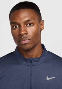 Nike navy blå zip-up jakke har en høj krave og et lys grå swoosh-logo på venstre bryst, lavet af et glat, åndbart materiale.