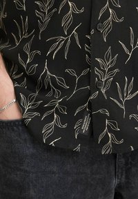 Zwarte shirt met een wit bladeren patroon, voorzien van een knoopsluiting en een binnenlabel; stof heeft een gestructureerde uitstraling, gecombineerd met donkere jeans.