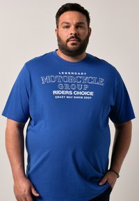 Modrá tričko s krátkým rukávem vyrobené z bavlny, se bílým potiskem textu: "LEGENDÁRNÍ MOTOCYKLOVÁ SKUPINA VOLBA JEZDCŮ ŠÍLENÝ ZPŮSOB OD ROKU 2007."