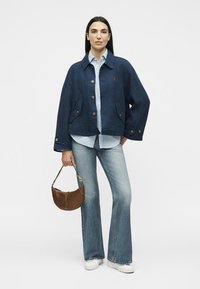 Γυναίκα που φοράει σκούρο μπλε σακάκι με κουμπιά, ανοιχτό μπλε πουκάμισο, καμπάνα τζιν, λευκά αθλητικά παπούτσια, κρατώντας καφέ τσάντα με κοντό λουράκι.