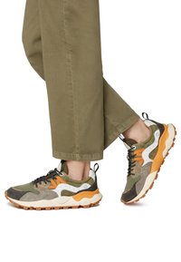 Scarpe sportive multicolori con una combinazione di verde, arancione e beige, caratterizzate da pannelli testurizzati. Suola robusta con scanalature profonde per una migliore trazione.