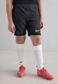 Mustad nailonnõudmispüksid, millel on kuldne Nike logo, ühendatud valgete põlvikute ja roosa jalgpallijalatsitega, millel on türkiissinised aktsendid.