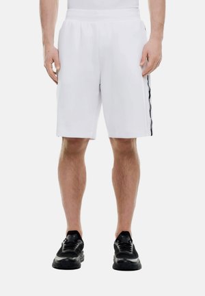 Hombre con pantalones cortos blancos hasta la rodilla con una franja lateral negra y zapatos deportivos negros, de pie sobre un fondo blanco.