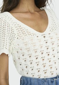 Top de punto blanco con escote en V y mangas cortas, que presenta un diseño de trabajo abierto con un patrón texturizado. Combinado con unos vaqueros de cintura alta.