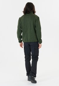 Giacca in pile verde con colletto alto, abbinata a pantaloni cargo blu. I pantaloni hanno tasche laterali e una vestibilità affusolata, con scarpe da trekking nere.