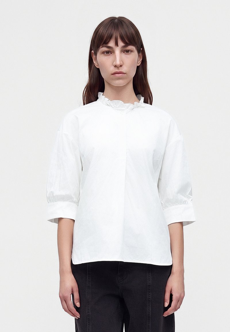 Femme aux cheveux bruns et raides portant un chemisier blanc à col volanté avec des manches trois-quarts bouffantes et un pantalon noir, debout devant un fond uni.