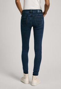 Jeans ajustados de color azul oscuro con cintura alta, con bolsillos traseros, una textura suave y un corte ceñido, combinados con zapatillas de color claro.
