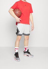 Person in red shirt and white basketball shorts holds basketball, wearing white socks with red design and black-and-white patterned basketball shoes.