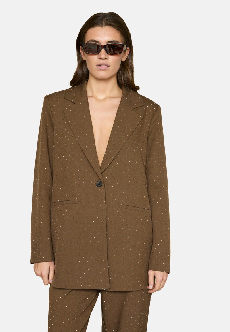 Brun blazer med et subtilt prikket mønster, enkeltknappslukning, to forlommer og skarpe revers. Brugtes over en neutral top.