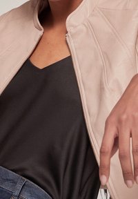 Světle růžová kožená bunda s prošívanými detaily, doplněná černým tričkem s výstřihem do V. Bunda má zipový uzávěr a přiléhavý střih.