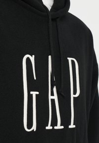 Čierna mikina s kapucňou s veľkým bielym logom GAP cez hruď a čiernymi šnúrkami visiace z kapucne.