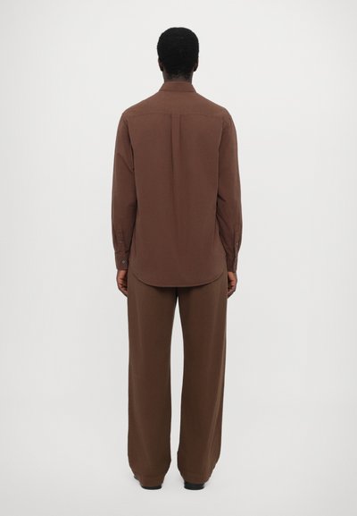 Chemise marron à manches longues avec col boutonné, ourlet droit et pli au dos, associée à un pantalon large assorti. Texture lisse.