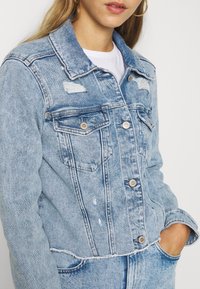 Light blue denim jacket with distressed detailing, button closures, chest pockets, and a frayed hem, featuring a collared design.