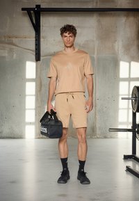 Tanosport set koji se sastoji od majice kratkih rukava i kratkih hlača, s Adidas logotipom. Crne sportske cipele i crna teretna torba upotpunjuju izgled.