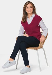 Diep V-hals, mouwloze, gebreide trui in bordeauxrood over een witte shirt, gecombineerd met donkere jeans en lichte sneakers, zittend op een geweven stoel.