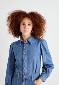 Macacão de ganga em azul médio com mangas bufantes, fecho frontal com botões e cintura ajustada. Possui bolsos de remendo e um acabamento texturizado.