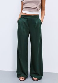 Frau trägt dunkelgrüne weite Hosen mit Falten und Taschen, kombiniert mit Sandalen und einem beigefarbenen cropped Top, lehnt gegen eine weiße Wand.