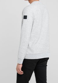 Šedá mikina vyrobená z texturované tkaniny, s kulatým výstřihem, žebrovanými manžetami a černou nášivkou na levém rukávu.