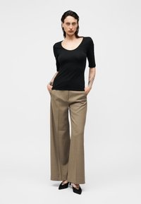 Vrouw met tatoeages die een zwart topje met ronde hals, beige wijde broek en zwarte puntige hakken draagt, staand met handen in de zakken.