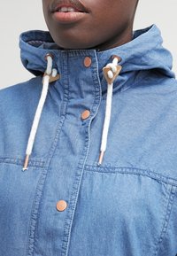 Jeansjacka i ljusblått med huva, med metallknappar, dragsko med läderdetaljer och en mjuk textur.