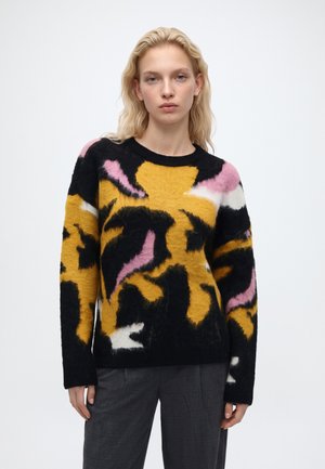 Femme blonde portant un pull doux avec de grands motifs abstraits jaunes, noirs, roses et blancs, et un pantalon gris foncé, debout sur un fond blanc.