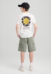 T-shirt bianco con una grande grafica di un fiore giallo e testo nero sul retro, abbinato a pantaloni cargo verdi e sneakers bianche.