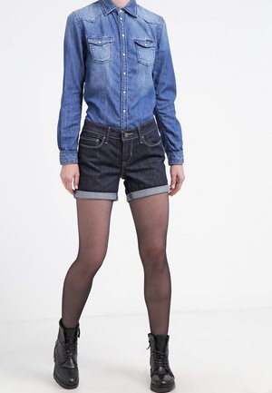 Persona che indossa una camicia di jeans con bottoni e tasche sul petto, pantaloncini di jeans scuri arrotolati, collant neri trasparenti e stivaletti neri con lacci.