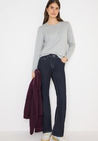 Maglia grigia a maniche lunghe, jeans svasati blu navy con cuciture a contrasto, tiene una giacca pelosa bordeaux, indossa sneaker bianche e blu.