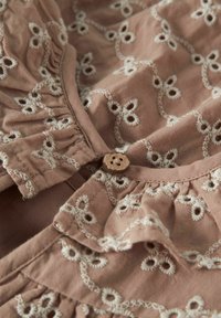 Gaiši rozā audums ar baltu izšūtu ziedu rakstu, volānu malām un koka pogas aizdari. Mīksta tekstūra un smalkas šūšanas detaļas.