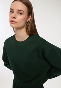 Zielony, oversize'owy sweter z okrągłym dekoltem, miękką fakturą i opadającymi ramionami. Posiada ribbowany kołnierz i mankiety dla większego komfortu.