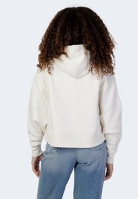 Hoodie bianco accorciato realizzato in tessuto morbido, con un ampio cappuccio, spalle scese e polsini a costine. Abbinato a jeans azzurri chiari.