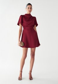 Bordo satino suknelė su trumpomis plazdančiomis rankovėmis, aptemptu liemeniu ir platėjančia sijonėle. Modelis laiko mažą, neutralios spalvos delninę ir dėvi juodus dirželius ant kulnų.